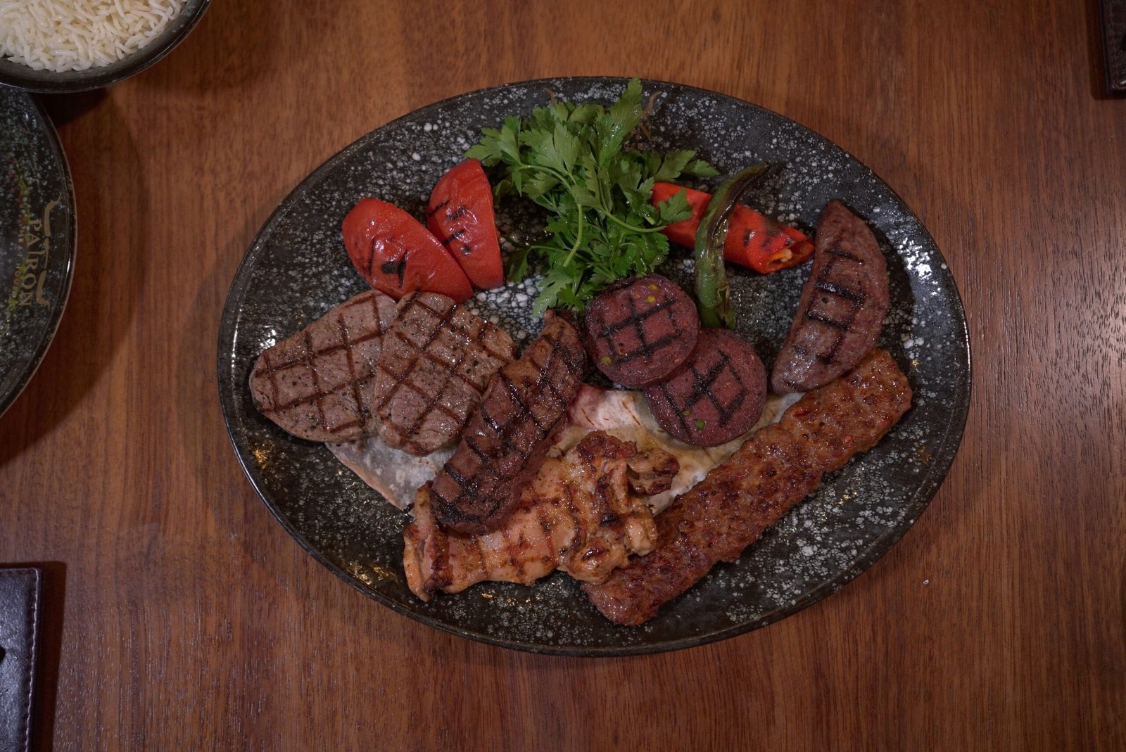 "Overhead view of a rustic plate of assorted grilled meats with charred tomatoes, green peppers and fresh parsley"